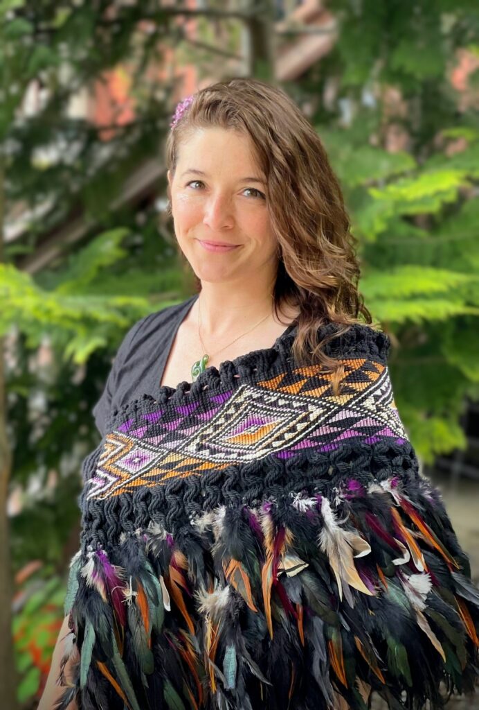 Image description: a white-bodied person with wavy brown hair falling past their shoulders, standing in portrait style, seen from the waist up. The person is smiling at the camera. They have a green jade pendant on a gold chain around their neck and are wearing a black, purple and orange coloured woven cloak, adorned with feathers of the same colour. The background is blurred with evergreen trees.